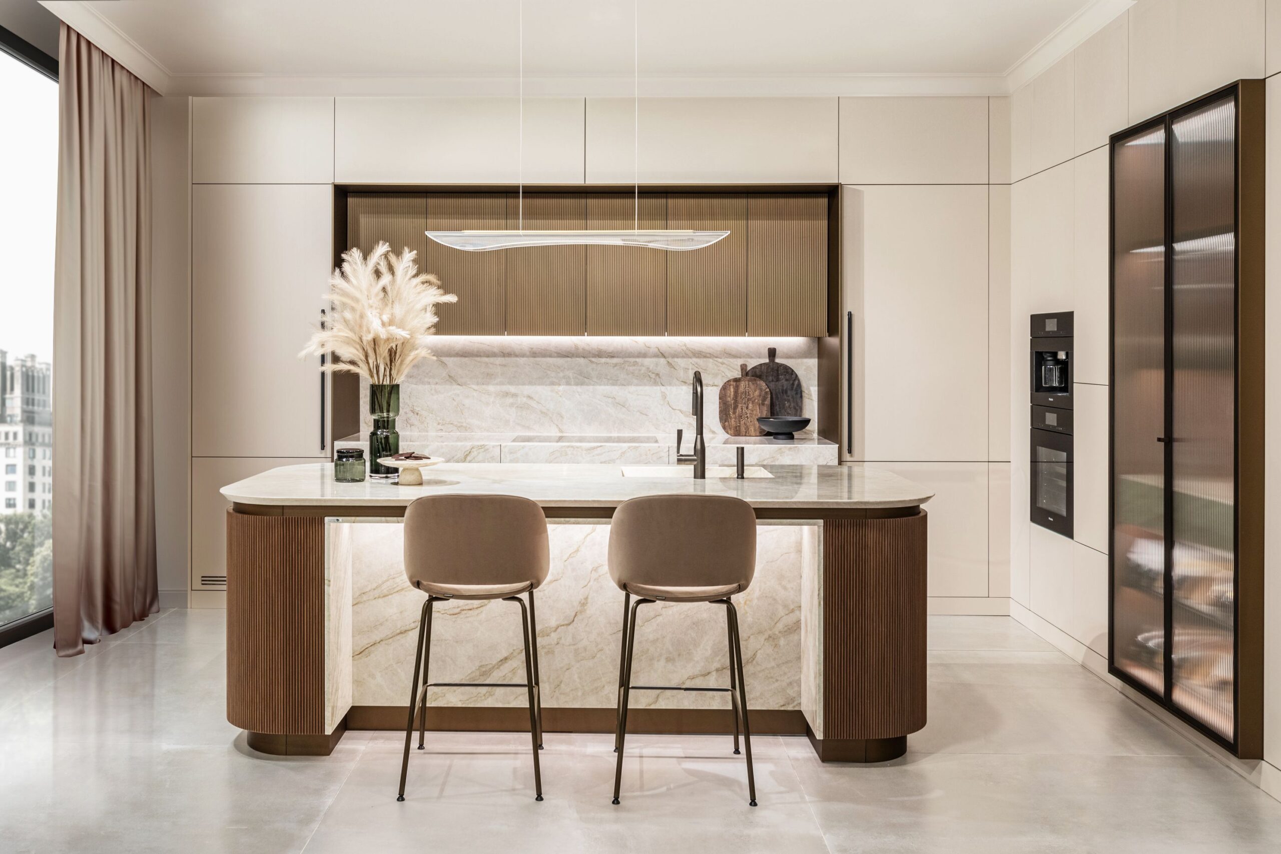 Modern kitchen with a marble island, two chairs, pendant light, and cabinets. Pampas grass decorates the island. Large windows with curtains in the background.