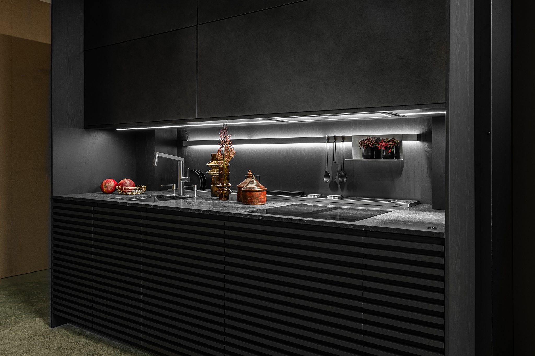 A modern kitchen with dark cabinetry, a marble countertop, and under-cabinet lighting. There are a few decorative items and a red pot on the counter, along with two pomegranates in a basket.