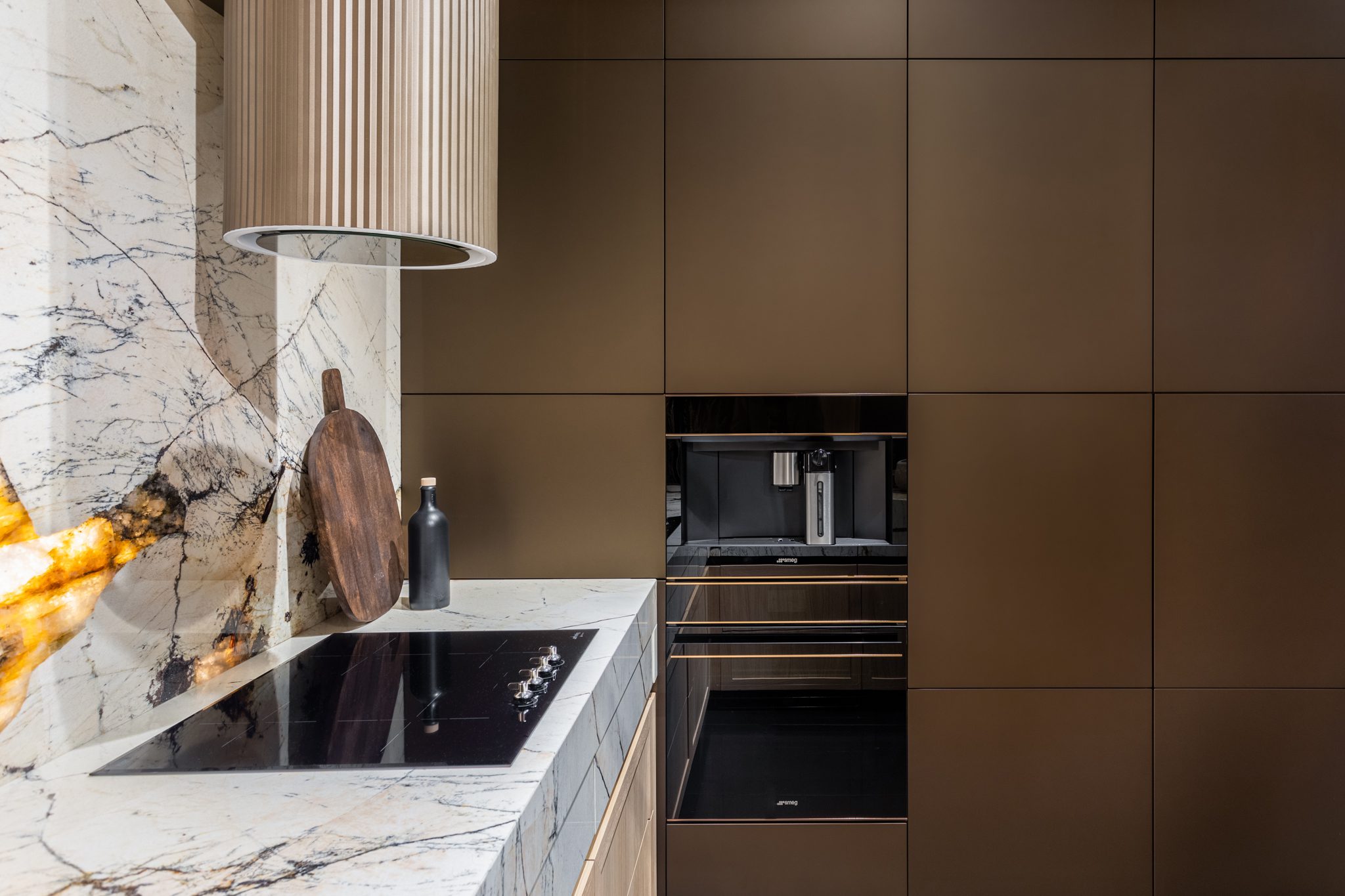 Modern kitchen with sleek marble countertops, a cooktop, wall-mounted oven, and dark paneled cabinets. A cutting board and bottle are placed on the counter.
